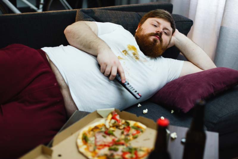 Udebljali čovek leži na kauču jede picu i gleda tv